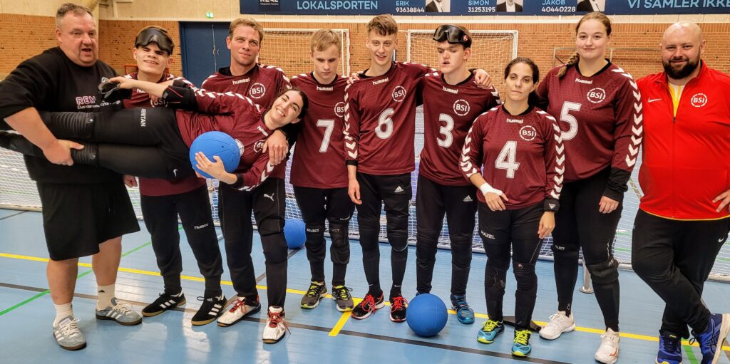 Gruppebillede af 10 mennesker i røde spillertrøjer og med bolde. En kvinde bliver båret af tre mænd.